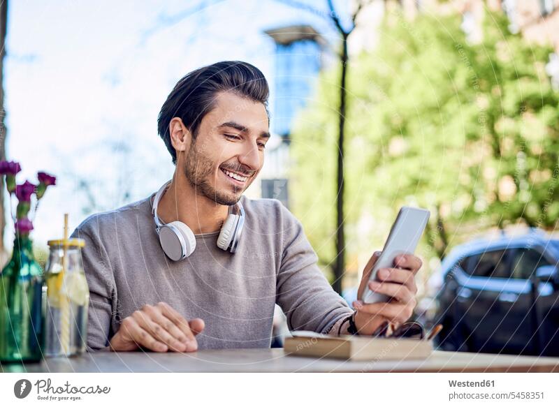 Glücklicher junger Mann, der ein Mobiltelefon in einem Café im Freien benutzt Handy Handies Handys Mobiltelefone Straßencafe Straßencafes Strassencafe