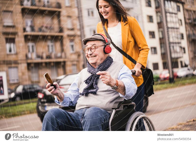Lachende junge Frau schiebt glücklichen älteren Mann mit Kopfhörern und Smartphone im Rollstuhl Leute Menschen People Person Personen Europäisch Kaukasier