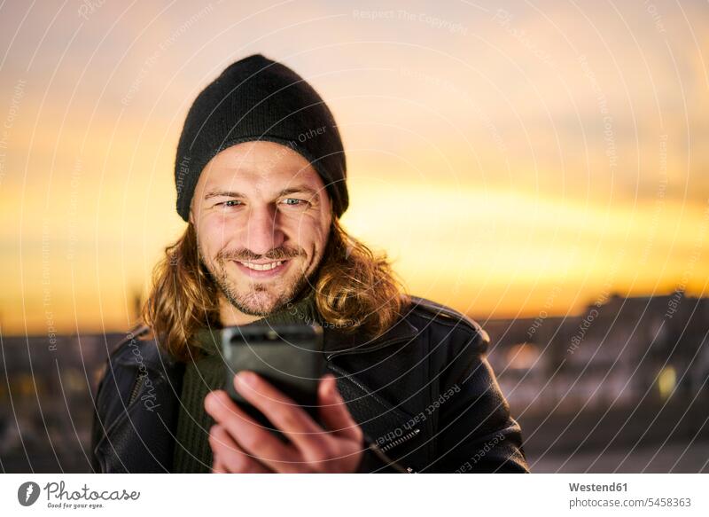 Porträt eines bärtigen jungen Mannes mit Wollmütze, der bei Sonnenuntergang auf sein Mobiltelefon schaut Smartphone iPhone Smartphones Portrait Porträts