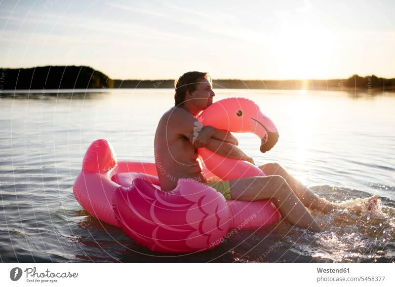 Junger Mann mit Flamingo-Pool schwimmt auf einem See entspannen relaxen sitzend sitzt Arm umlegen Umarmung Umarmungen abends Jahreszeiten sommerlich Sommerzeit