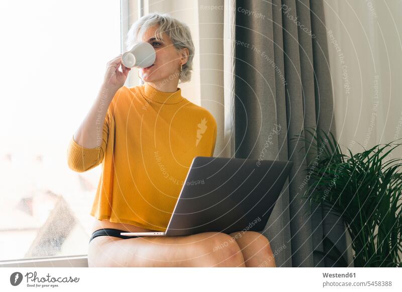Frau sitzt am Fenster zu Hause mit Laptop trinken Kaffee aus Tasse Leute Menschen People Person Personen Alleinstehende Alleinstehender Singles Unverheiratete