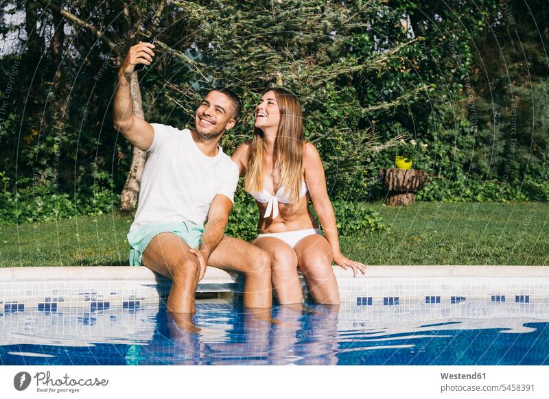 Lächelnder Mann, der ein Selfie mit seiner Freundin über ein Smartphone macht, während er am Pool gegen Bäume sitzt Farbaufnahme Farbe Farbfoto Farbphoto
