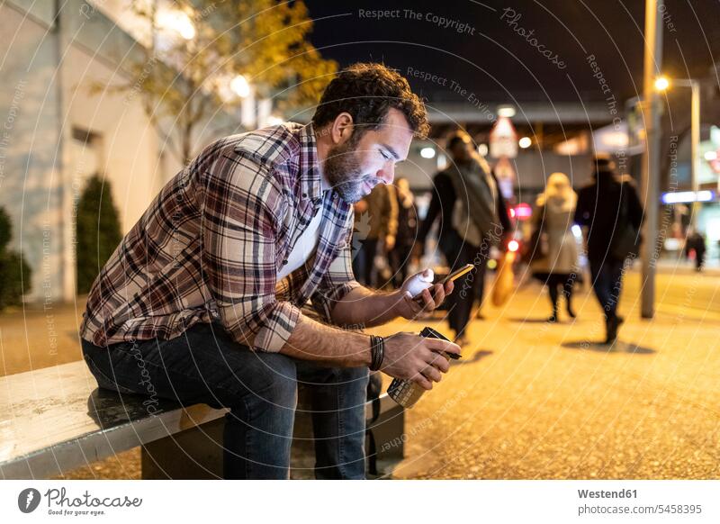 UK, London, lächelnder Mann sitzt auf einer Bank und schaut auf sein Telefon bei Nacht Männer männlich Sitzbänke Bänke Sitzbank Handy Mobiltelefon Handies