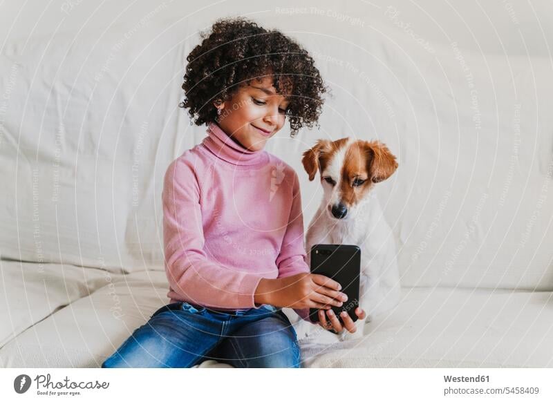 Porträt eines lächelnden kleinen Mädchens, das mit ihrem Hund auf der Couch sitzt und ein Selfie mit dem Smartphone macht Leute Menschen People Person Personen