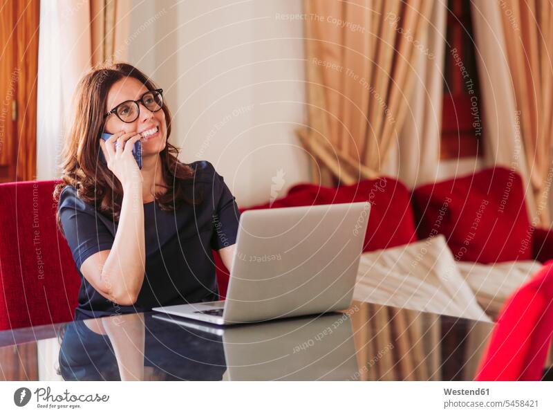 Lächelnde Geschäftsfrau telefoniert mit dem Handy, während sie mit dem Laptop am Tisch im Wohnzimmer sitzt Farbaufnahme Farbe Farbfoto Farbphoto Spanien