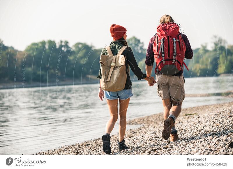 Rückansicht eines jungen Paares mit Rucksäcken, das Hand in Hand am Flussufer geht Rucksack gehen gehend Fluesse Fluß Flüsse Pärchen Partnerschaft Gewässer