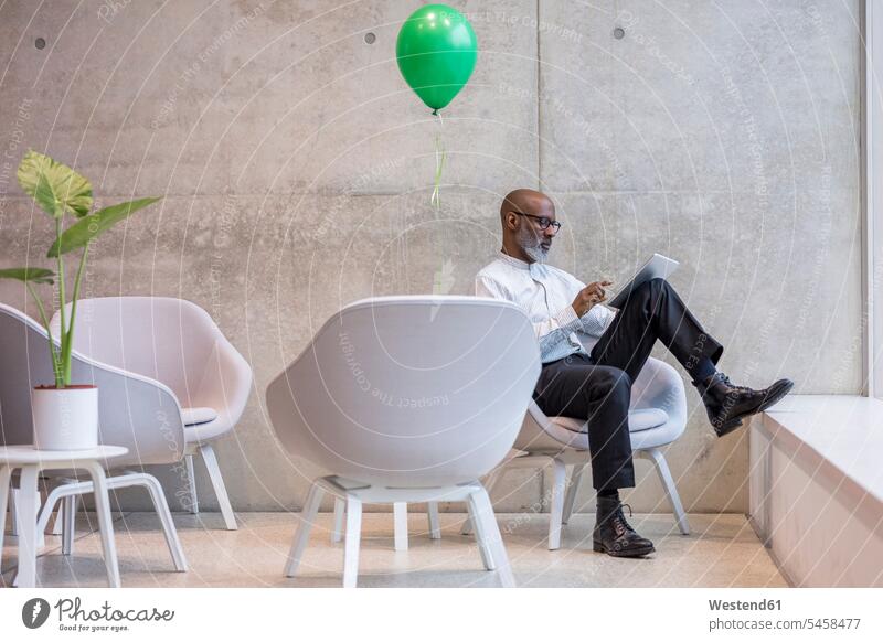 Älterer Geschäftsmann mit grünem Ballon sitzt auf einem Sessel und benutzt ein digitales Tablet Businessmann Businessmänner Geschäftsmänner benutzen benützen
