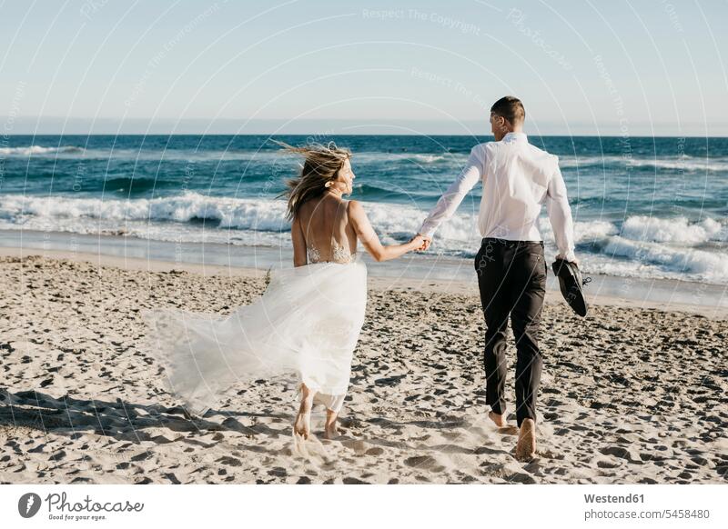 Rückansicht von Braut und Bräutigam beim Laufen am Strand Feiern Fest Feste Festlichkeit Festlichkeiten Heirat heiraten Hochzeiten Bräute Bräutigame Kleider