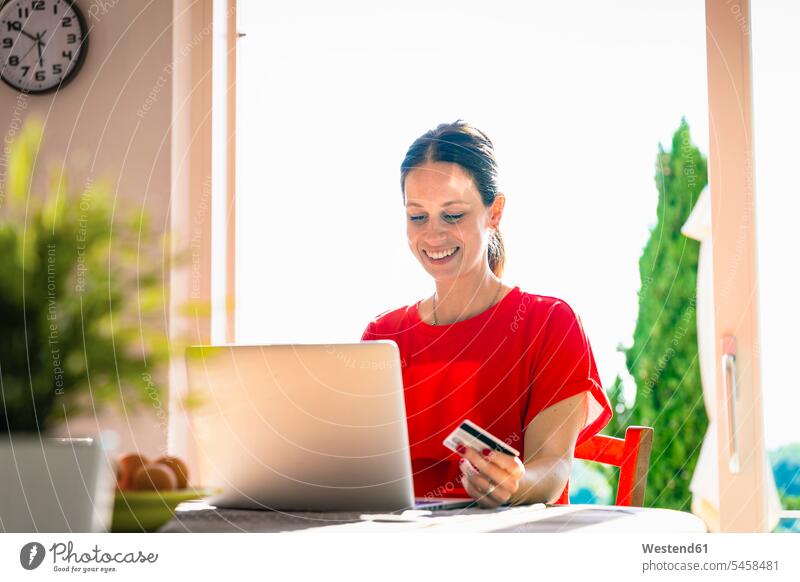 Lächelnde schöne Frau mit Kreditkarte, während sie den Laptop am Esstisch gegen das Fenster hält Farbaufnahme Farbe Farbfoto Farbphoto Innenaufnahme