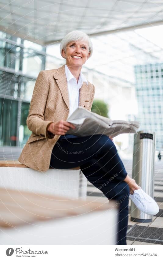 Lächelnde ältere Geschäftsfrau sitzt in der Stadt mit Zeitung Geschäftsfrauen Businesswomen Businessfrauen Businesswoman staedtisch städtisch Frau weiblich