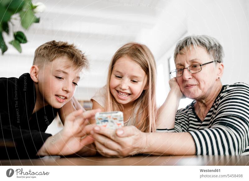 Glückliche Großmutter und Enkelkinder bei der Nutzung eines Mobiltelefons zu Hause Wissen Tisch Tische erklären erklaeren spielen Häusliches Leben Wohnen