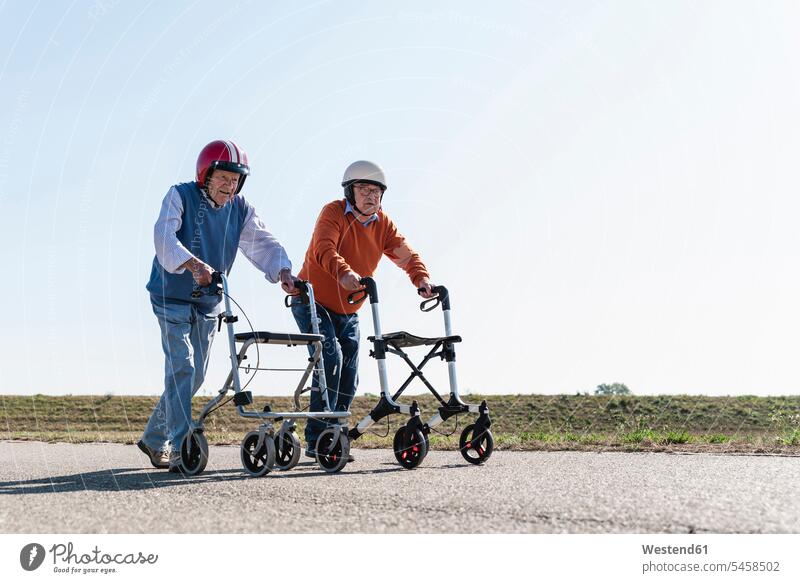 Zwei alte Freunde mit Schutzhelmen, die an einem Rennen mit Rollatoren teilnehmen Rollgestell Gehhilfe Rollwagen laufen rennen Wettkampf Wettkaempfe Wettstreit