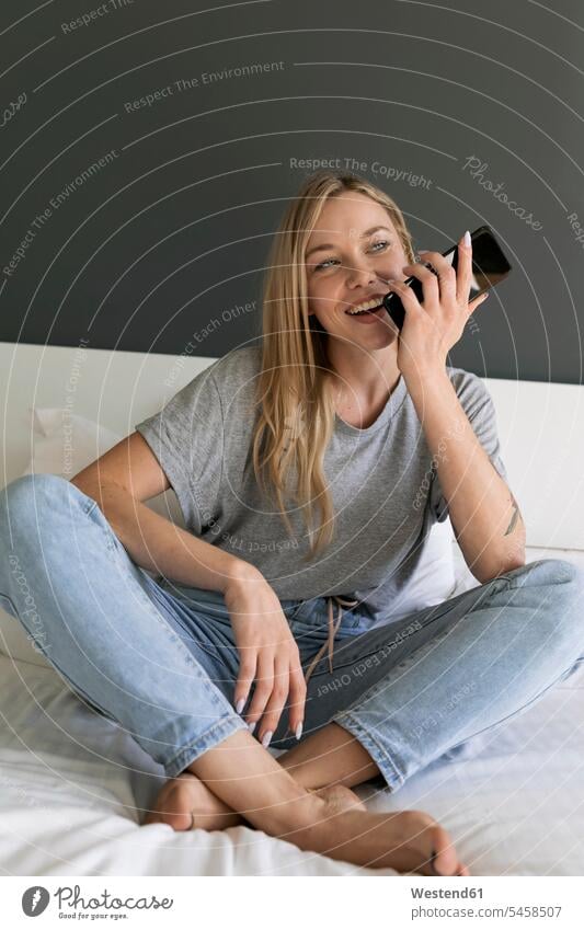 Glückliche junge Frau sitzt auf dem Bett und benutzt ein Mobiltelefon sitzen sitzend glücklich glücklich sein glücklichsein weiblich Frauen Handy Handies Handys
