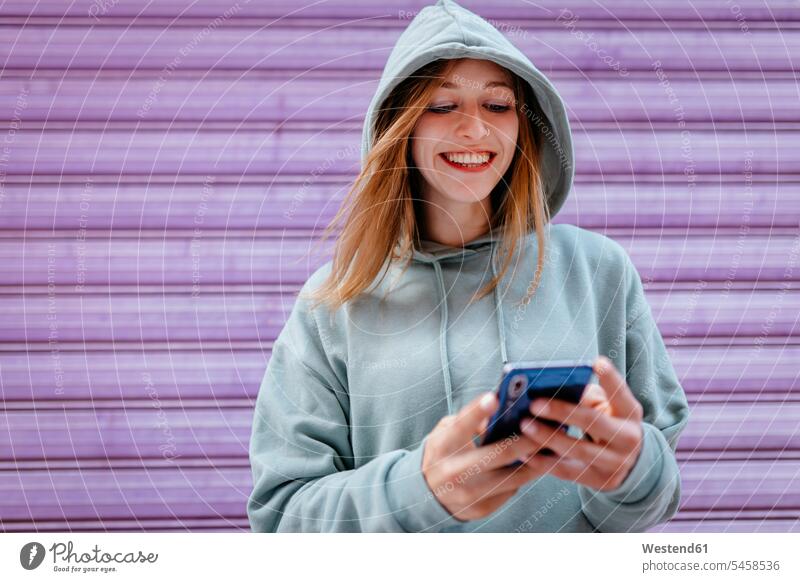 Porträt einer jungen blonden Frau mit grünem Kapuzenpulli und lila Hintergrund Telekommunikation telefonieren Handies Handys Mobiltelefon Mobiltelefone Lektüre