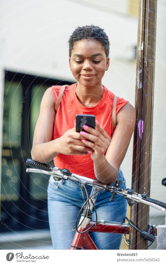 Porträt einer lächelnden jungen Frau, die sich auf den Lenker eines Fahrrads stützt und auf ihr Handy schaut Leute Menschen People Person Personen Afrikanisch