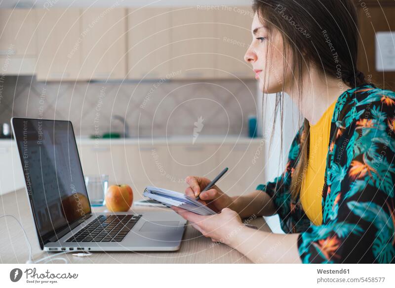 Junge Frau sitzt zu Hause am Tisch mit Laptop und macht sich Notizen Notebook Laptops Notebooks Tische notieren weiblich Frauen Zuhause daheim Computer Rechner