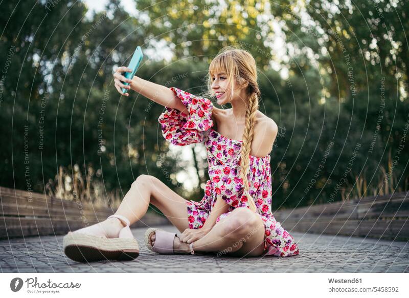 Porträt einer lächelnden jungen Frau, die im Sommer auf der Strandpromenade sitzt und ein Selfie mit ihrem Smartphone macht Handies Handys Mobiltelefon