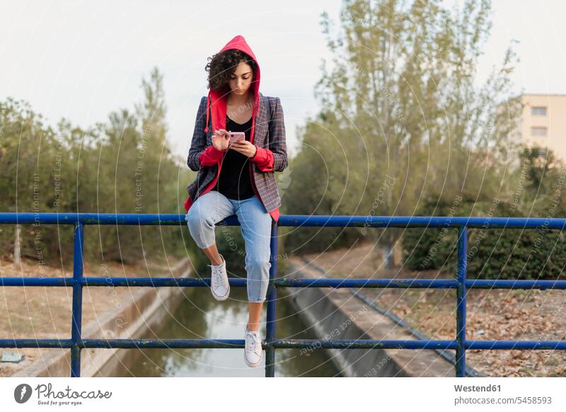 Junge Frau sitzt auf einem Geländer und benutzt ein Mobiltelefon Leute Menschen People Person Personen Europäisch Kaukasier kaukasisch 1 Ein ein Mensch