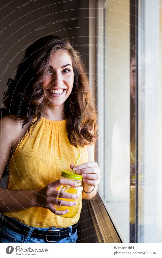 Schöne junge Frau hält Maurerglas mit Saft in der Hand, während sie zu Hause am Fenster lächelt Farbaufnahme Farbe Farbfoto Farbphoto Spanien