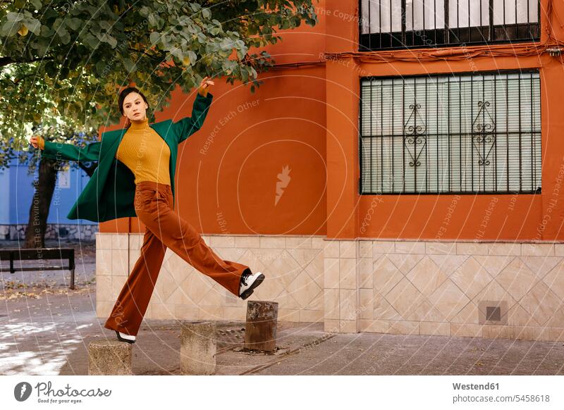 Schöne Frau in grüner Jacke klettert in der Stadt auf einen Poller Farbaufnahme Farbe Farbfoto Farbphoto Spanien Freizeitkleidung Freizeitbekleidung casual