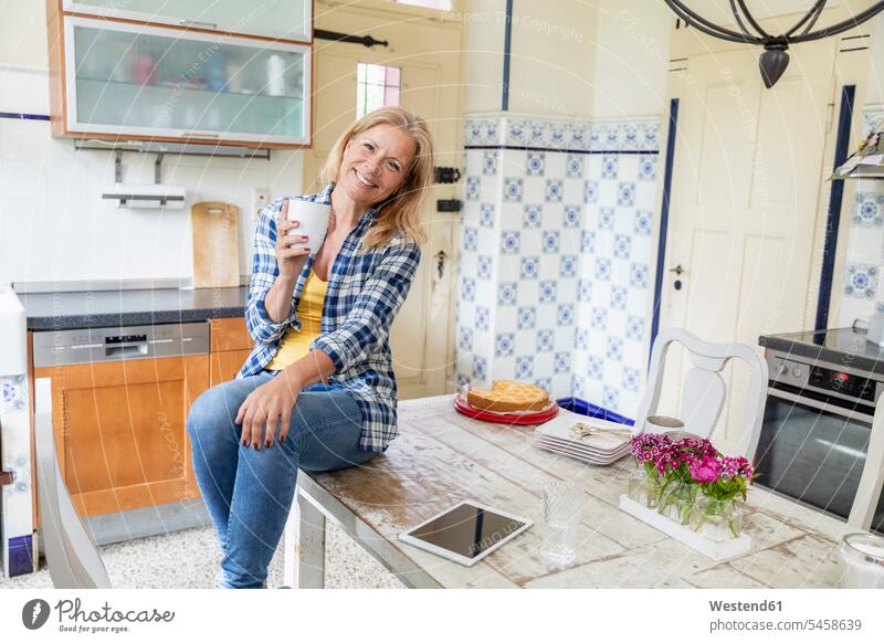 Porträt der lächelnden reife Frau mit Kaffeetasse sitzt auf Esstisch in der Küche Leute Menschen People Person Personen Europäisch Kaukasier kaukasisch 1 Ein