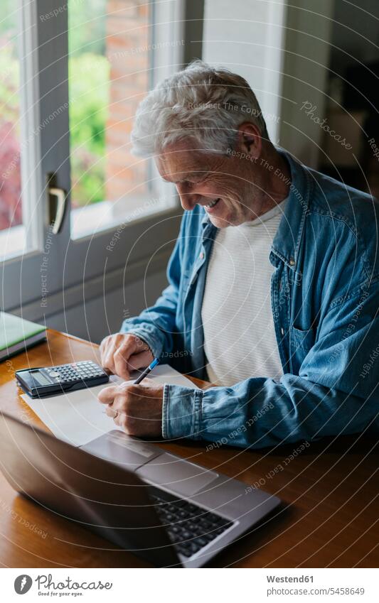 Glücklicher älterer Mann schreibt auf Papier, während er zu Hause Taschenrechner und Laptop benutzt Farbaufnahme Farbe Farbfoto Farbphoto Innenaufnahme