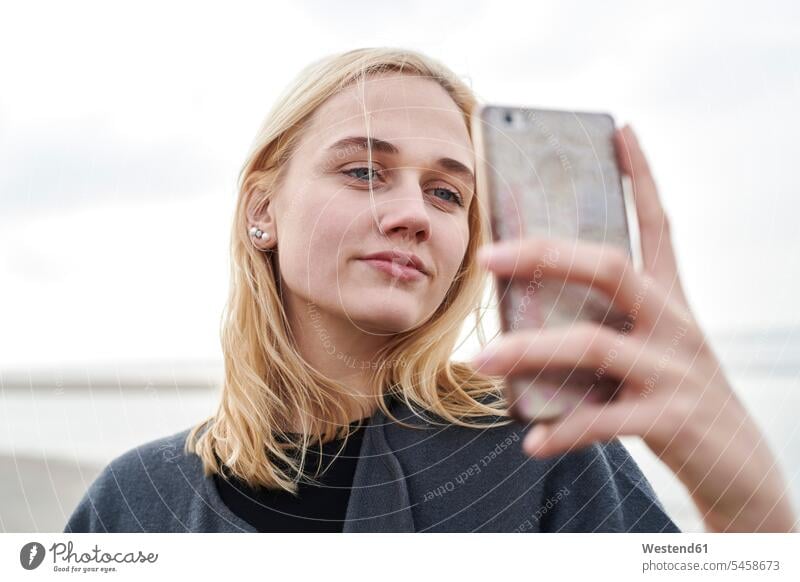 Porträt einer blonden jungen Frau, die ein Selfie mit ihrem Smartphone am Strand macht Portrait Porträts Portraits Selfies blonde Haare blondes Haar weiblich