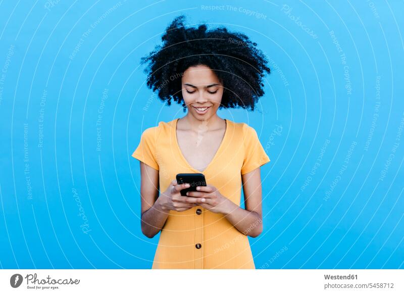 Lächelnde junge Frau benutzt Smartphone, während sie an blauer Wand steht Farbaufnahme Farbe Farbfoto Farbphoto Außenaufnahme außen draußen im Freien Tag