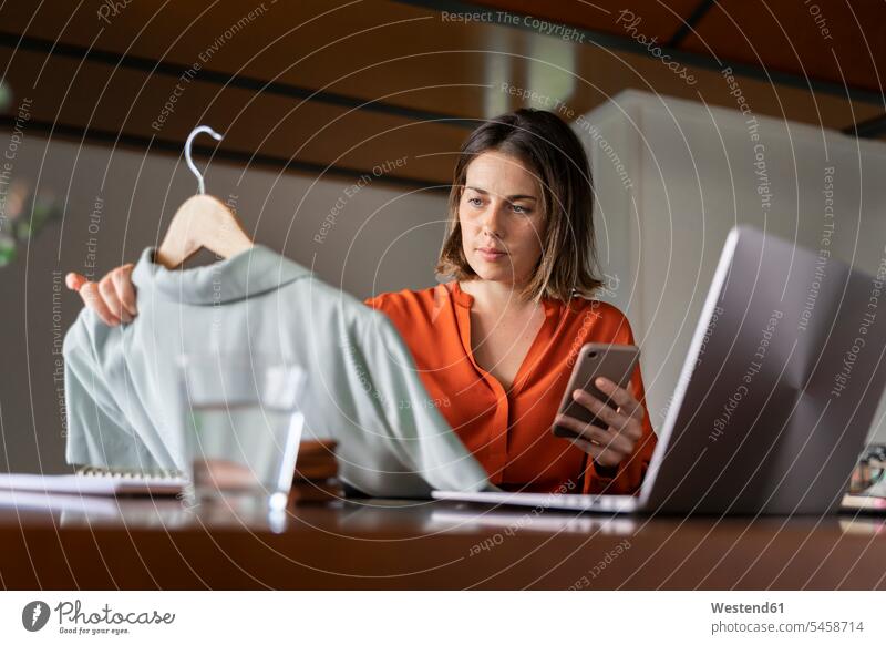 Geschäftsfrau mit Smartphone betrachtet Kleidung, während sie zu Hause sitzt Farbaufnahme Farbe Farbfoto Farbphoto Innenaufnahme Innenaufnahmen innen drinnen