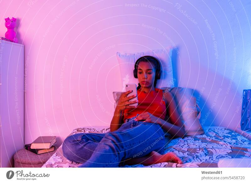 Teenager-Mädchen liegt im Bett und benutzt ein Smartphone, um zu Hause über Kopfhörer Musik zu hören Farbaufnahme Farbe Farbfoto Farbphoto Innenaufnahme