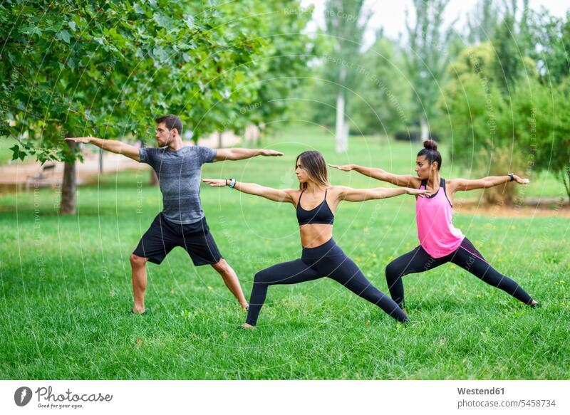 Junge Menschen üben Yoga in einem Park trainieren strecken dehnen Kriegerstellung Virabhadrasana Krieger-Stellung Parkanlagen Parks Fitness Gesundheit gesund