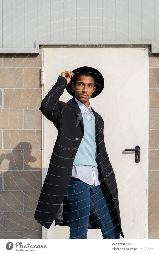 Modischer junger Mann hält Hut, während er an einem sonnigen Tag vor der Tür steht Farbaufnahme Farbe Farbfoto Farbphoto Außenaufnahme außen draußen im Freien