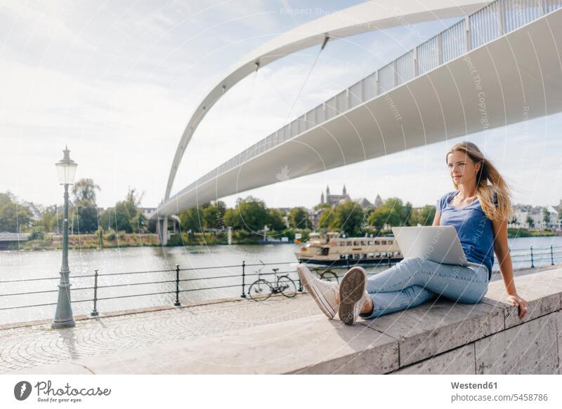Niederlande, Maastricht, junge Frau sitzt mit Laptop auf einer Mauer am Flussufer Notebook Laptops Notebooks Mauern sitzen sitzend weiblich Frauen Fluesse Fluß