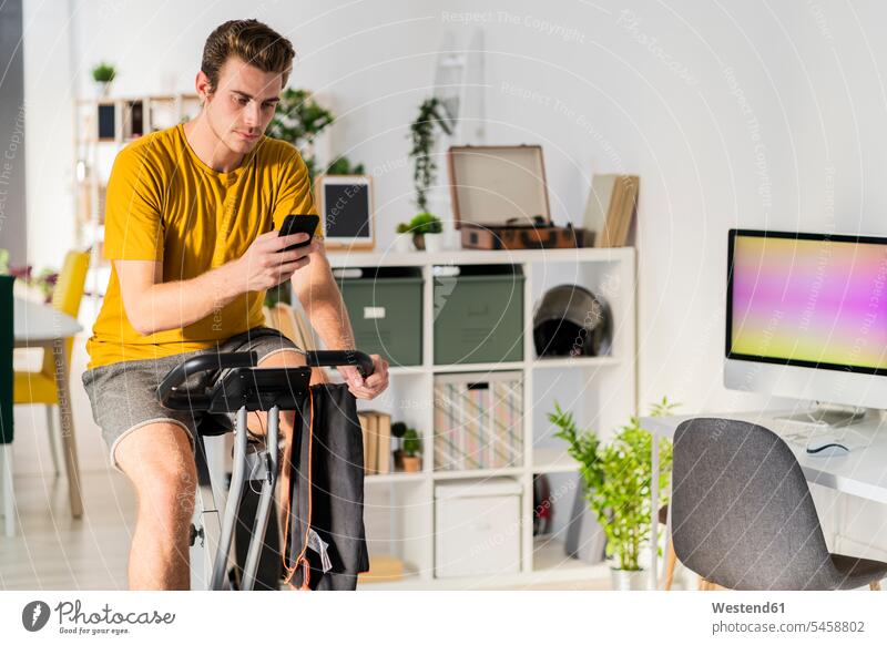 Mann benutzt Mobiltelefon, während er zu Hause mit Trainingsgeräten Rad fährt Farbaufnahme Farbe Farbfoto Farbphoto Innenaufnahme Innenaufnahmen innen drinnen