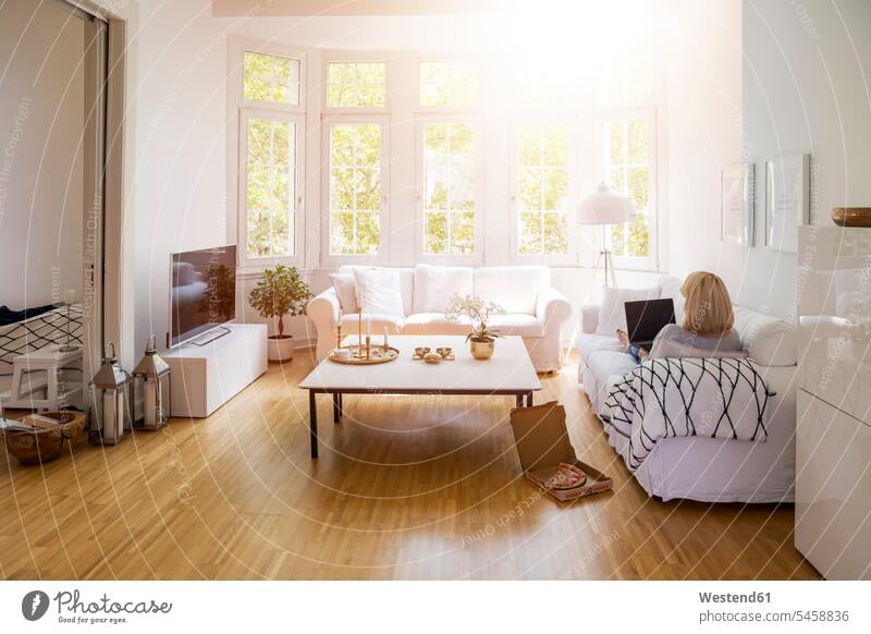 Rückenansicht einer blonden, reifen Frau, die im Wohnzimmer auf der Couch sitzt und einen Laptop benutzt Wohnraum Wohnung Wohnen Wohnräume Wohnungen Sofa