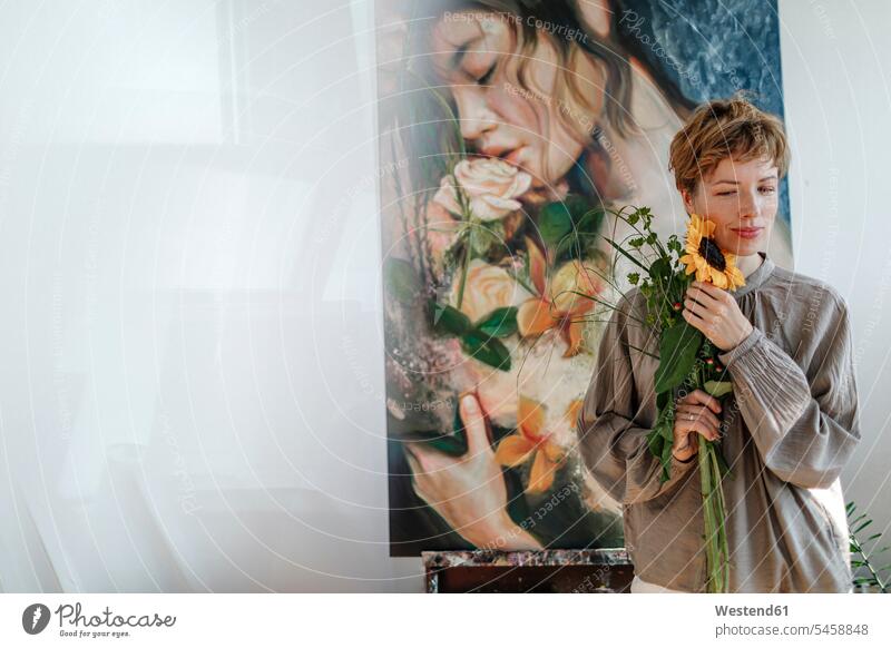 Nachdenkliche mittel-erwachsene Künstlerin, die eine Sonnenblume hält, während sie im Atelier gegen ein Kunstwerk steht Farbaufnahme Farbe Farbfoto Farbphoto