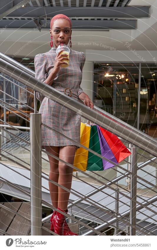 Junge Frau mit kurzen Haaren, die bunte Einkaufstaschen hält und auf der Treppe einen Smoothie trinkt Taschen Tuete Tueten Tüten Einkaufstüten Kleider Kauf