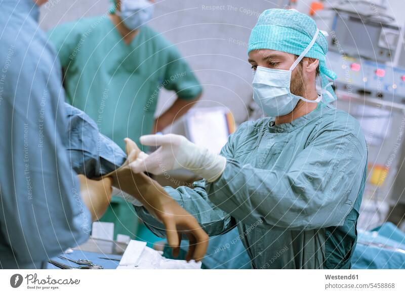 Assistentin, die dem Chirurgen hilft, vor einer Operation Handschuhe anzuziehen überziehen anziehen OP Operationen operieren Chirurgie Operationskittel helfen