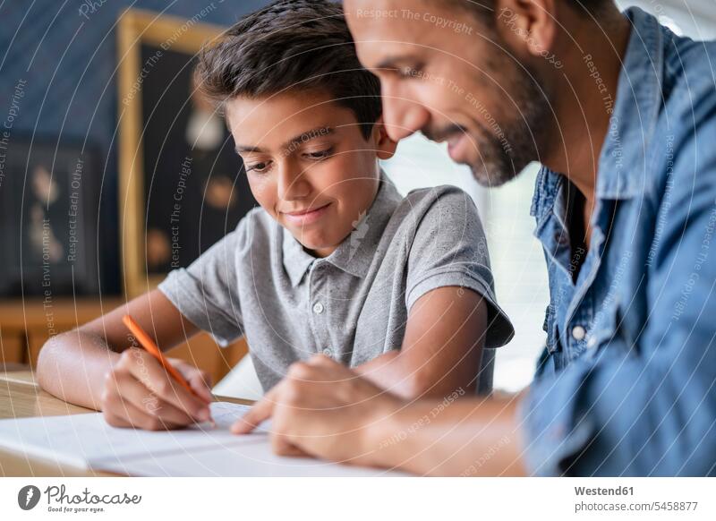 Lächelnder Vater hilft Sohn bei den Hausaufgaben Schüler Tisch Stift lernen lächeln schreiben sitzen Freude Zufriedenheit Zuhause Leistung Erfolg Gemeinsam