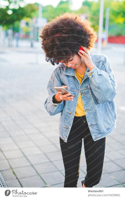 Junge Frau mit Afrofrisur benutzt Smartphone und Kopfhörer in der Stadt Leute Menschen People Person Personen gelockt gelockte Haare gelocktes Haar lockig
