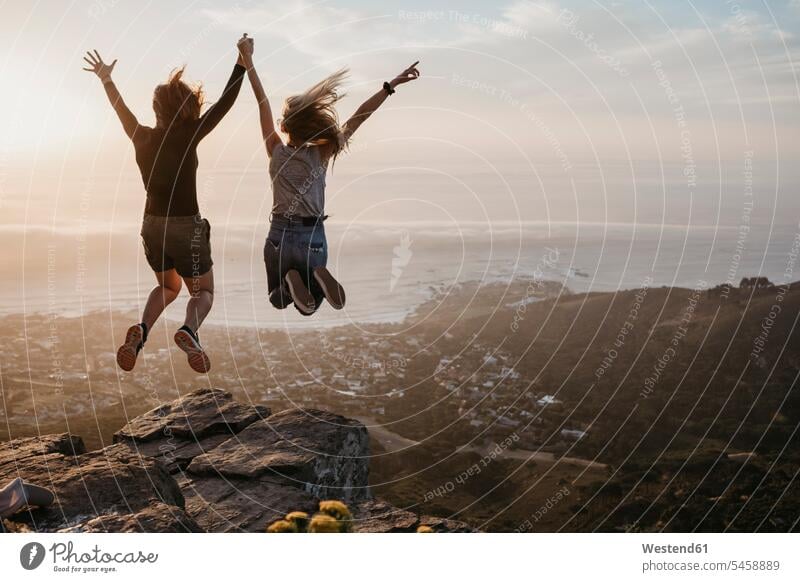 Südafrika, Kapstadt, Kloof Nek, zwei Frauen springen bei Sonnenuntergang auf Felsen Freundin hüpfen weiblich Sonnenuntergänge Gestein Steine Sprung Spruenge