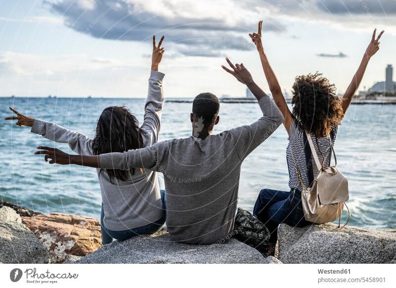 Drei Freunde sitzen am Strand, schauen auf das Meer, jubelnd, Rückansicht Frieden friedlich Vergnügen genießen freuen Amüsement Freude vergnügt amusieren