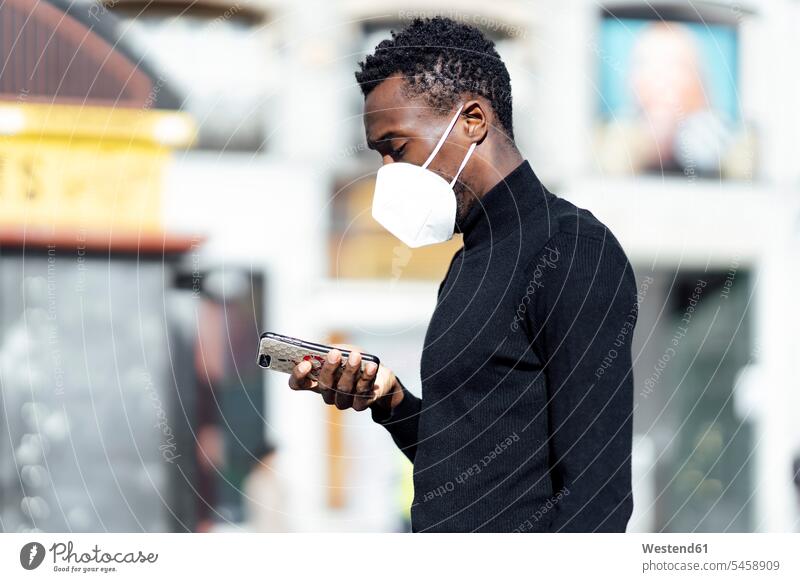 Junger Mann mit Gesichtsmaske, der ein Mobiltelefon benutzt, während er im Freien steht Farbaufnahme Farbe Farbfoto Farbphoto Außenaufnahme außen draußen Tag