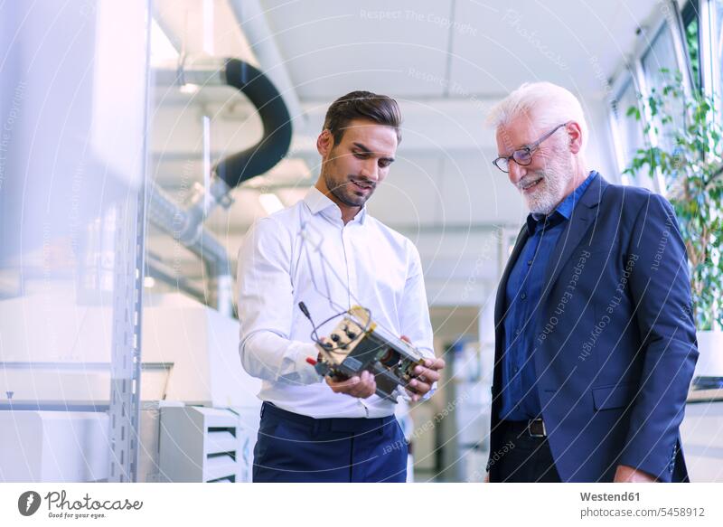 Junger männlicher Ingenieur hält Maschinenteil in der Hand, während er mit lächelndem Senior Manager in der Fabrik diskutiert Farbaufnahme Farbe Farbfoto