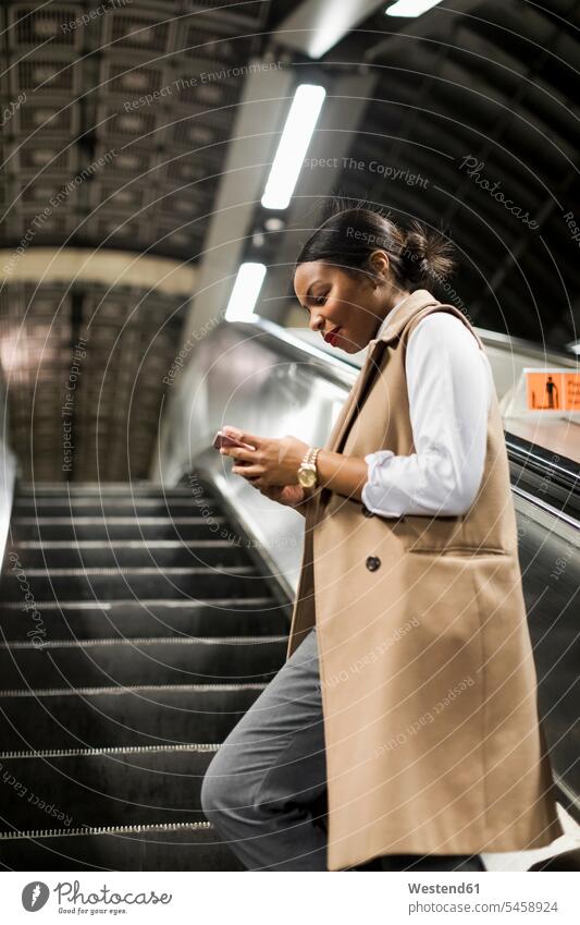 Großbritannien, London, lächelnde Geschäftsfrau steht auf der Rolltreppe der U-Bahn-Station und schaut auf ihr Handy Geschäftsfrauen Businesswomen