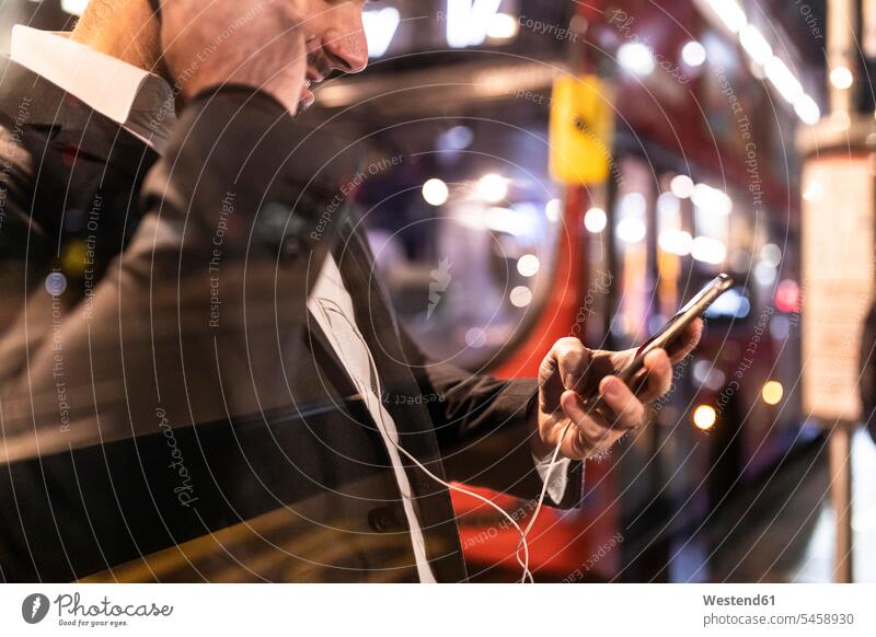 UK, London, Geschäftsmann mit Handy und Ohrstöpsel am Busbahnhof bei Nacht Bushaltestelle Haltestelle Haltestellen Bushaltestellen checken Businessmann