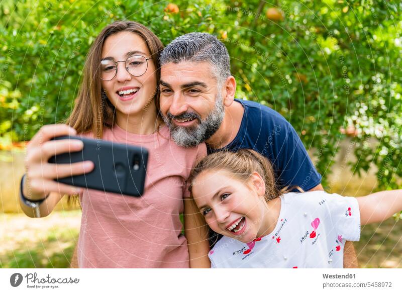 Glücklicher Vater mit zwei Töchtern macht Selfie im Garten T-Shirts Telekommunikation telefonieren Handies Handys Mobiltelefon Mobiltelefone Brillen entspannen