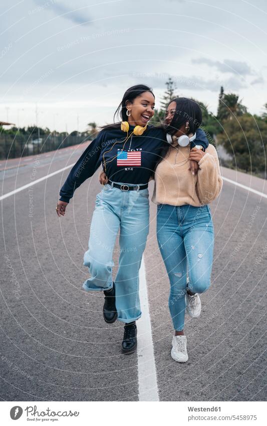 Zwei glückliche junge Frauen gehen auf einer Straße Freunde Kameradschaft Freundin Hosen Jeanshose Kopfhoerer gehend geht reden Arm umlegen Umarmung Umarmungen