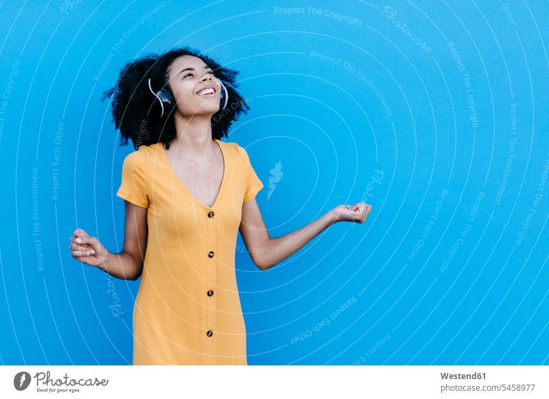 Glückliche junge Afro-Frau genießt es, Musik über Kopfhörer an blauer Wand zu hören Farbaufnahme Farbe Farbfoto Farbphoto Außenaufnahme außen draußen im Freien