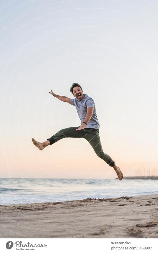 Glücklicher junger Mann springt bei Sonnenuntergang am Strand in die Luft Sonnenuntergänge Beach Straende Strände Beaches Männer männlich springen hüpfen